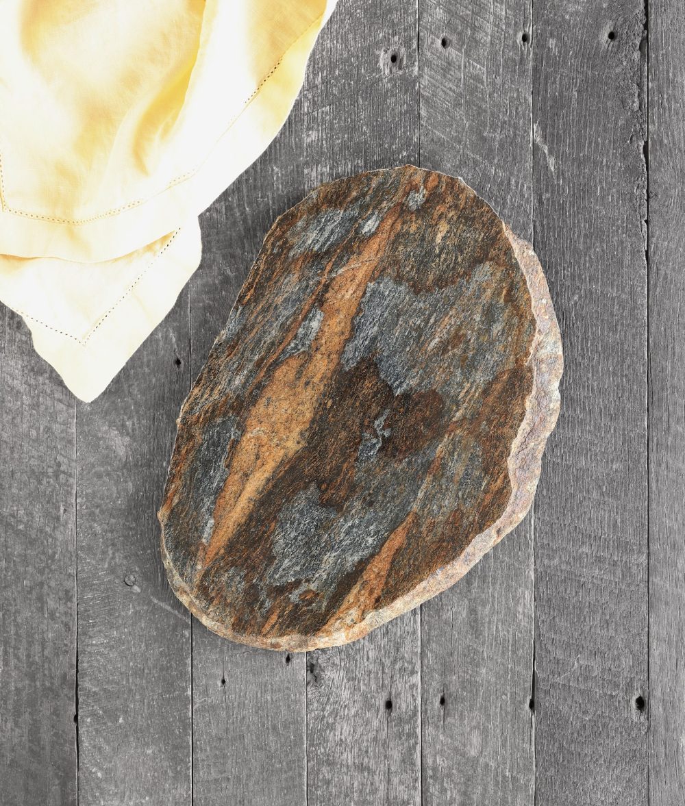looking down from above onto polished stone charcuterie board on gray wood background with yellow napkin in the corner. Stone has brown tones with orange veins, dark brown clusters, and visible portions of the live edge.